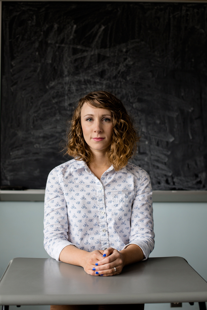 editorial photographer - a young teacher sits at a classroom desk in front of a chalk board